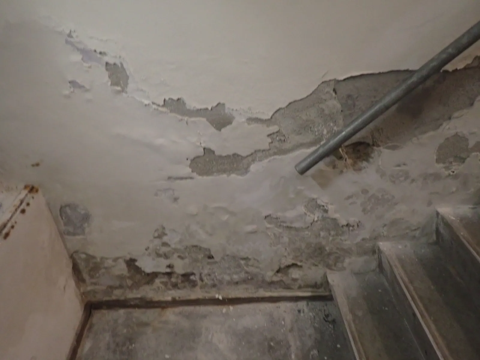 Overzicht van een kelder met vochtschade rond plafond, leidingwerk en trap